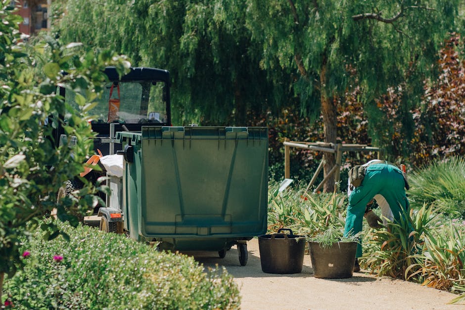 A gardener in teal work overalls is bent over, tending to plants in a well-maintained garden with a variety of shrubs, leafy plants, and small trees. The garden features a paved pathway bordered by lu