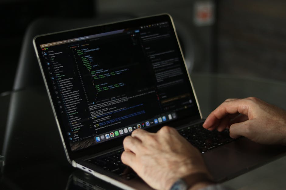 Close-up of a person's hands operating a laptop on a dark outdoor table, with the screen displaying lines of computer code in a text editor. The individual appears to be engaged in programming or web